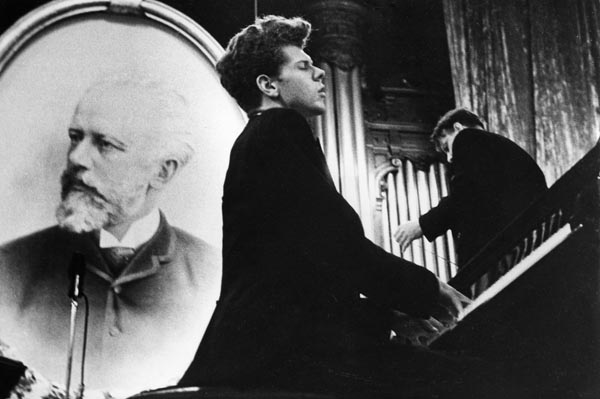 A black-suited Van Cliburn sits at a piano, playing with his eyes closed. A portrait of Tchaikovsky is propped behind him