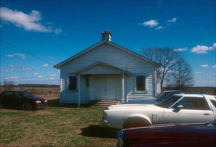 Under blue skies, a white clapboard church sits in a grassy field. 1970s cars are parked outside