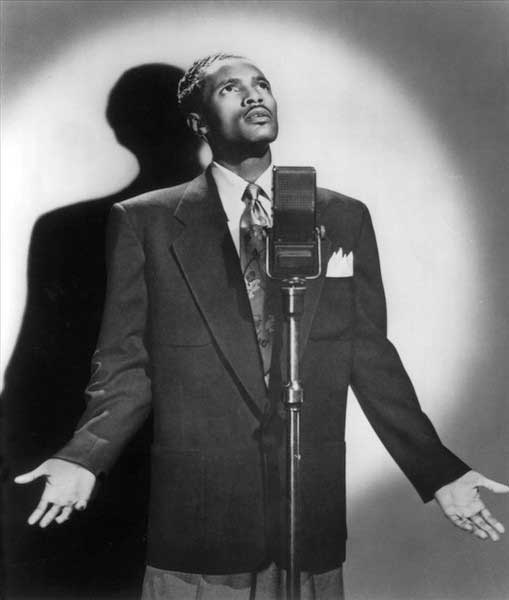 Percy is lit by a spotlight and stands before a microphone; with hands and arms extended, he looks up to the sky, as if singing to God