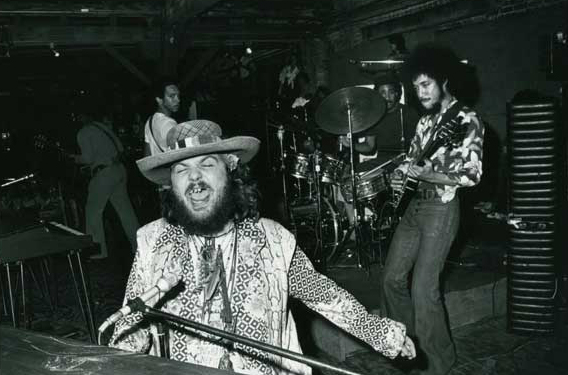 Dr. John sits at a piano singing enthusiastically while a man plays guitar behind him