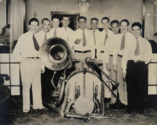 A group of men in white shirts and tie gather around a drum set