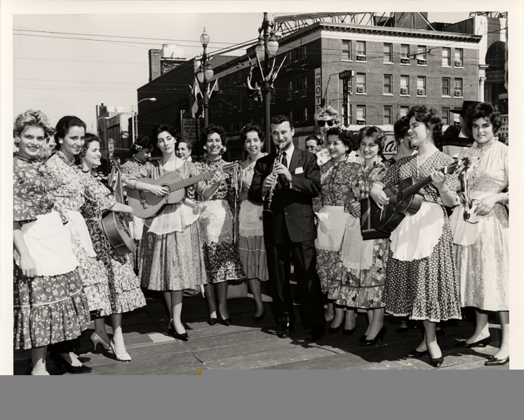 A group of women in flowered dresses stand around a be0suited Pete. Two of the women hold a guitar. They all pose on a street corner