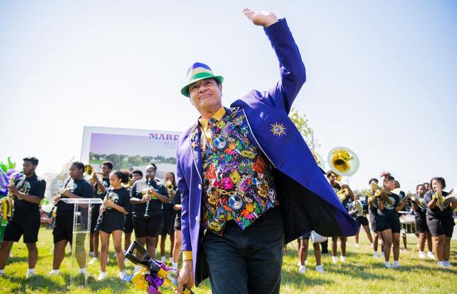 A person dressed in a festive blazer and hat waves to a crowd celebrating Mardi Gras.