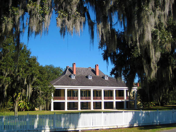 Front view of the Destrehan Plantation main house with white columns and moss-draped trees, surrounded by a white wooden fence.
