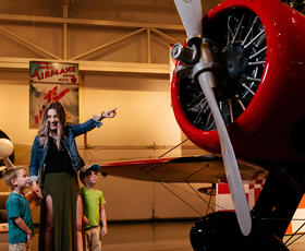 A woman with two children pointing at a large red airplane and its propeller inside a museum