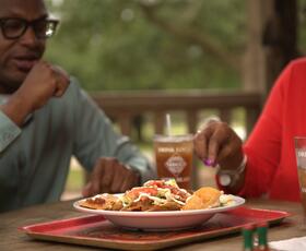 Two people share a plate of nachos at an outdoor table with iced drinks branded with "TABASCO" and "Avery Island."