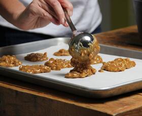 A person spoons fresh praline candy onto a baking tray lined with parchment paper.