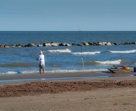 A fisherman casts a line from the beach at Grand Isle State Park with gentle waves rolling in toward a rock breakwater.