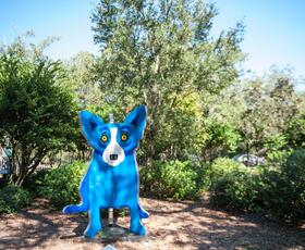 A bright blue dog sculpture stands along a wooded path in the Besthoff Sculpture Garden at City Park, surrounded by trees and landscaped greenery.