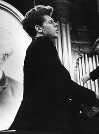 A black-suited Van Cliburn sits at a piano, playing with his eyes closed. A portrait of Tchaikovsky is propped behind him