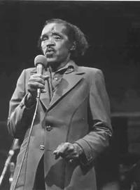 An older Percy wears and dark leisure suit and croons into a mic