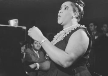 Nellie, in pearls and a black dress, hair in a bun, sings before a gathered audience