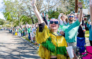 A person dressed in a gold and green fringe jacket waves at an oncoming parade float.